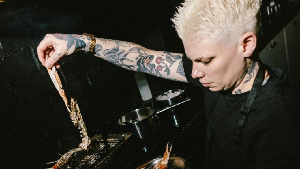 A woman with tattoos, short blonde hair and a black top holding up a piece of food