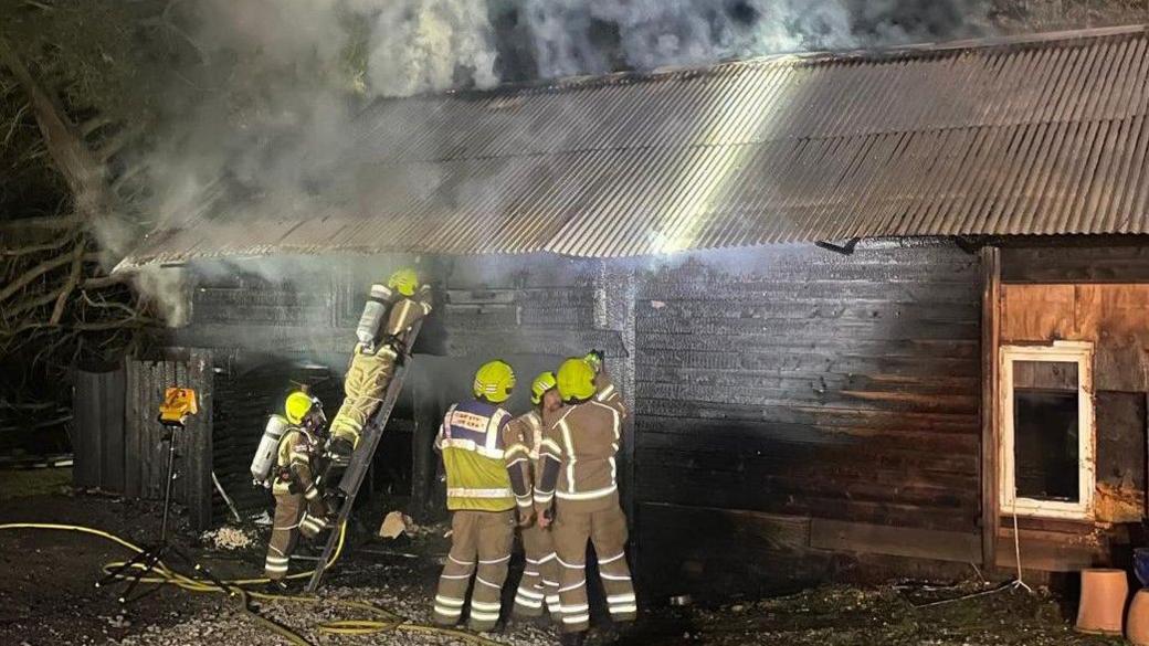 A group of firefighters work to extinguish a fire at an outbuilding. Three of the firefighters appear to be in discussion, while another is climbing a ladder to tackle the blaze. Smoke is rising from the structure.