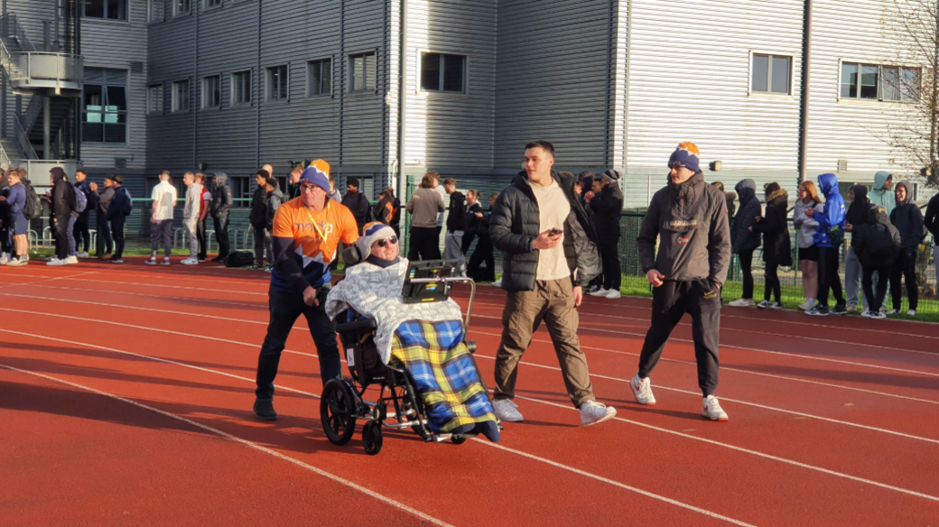 A man in a wheelchair is pushed around a running track by a man in an orange hat while two other men walk beside him. A crowd watches from the outside of the track.