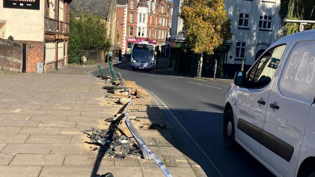 Bollards knocked down on the pavement on Fye Bridge Street, with the Mischief pub and the start of Magdalen Street visible in background. There is  a bus and white van on the road.