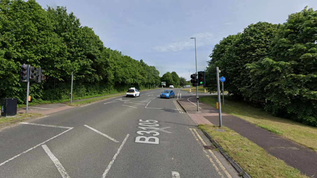 A B-road on a sunny day with traffic lights, cars, street markings and signage. Trees in the background.