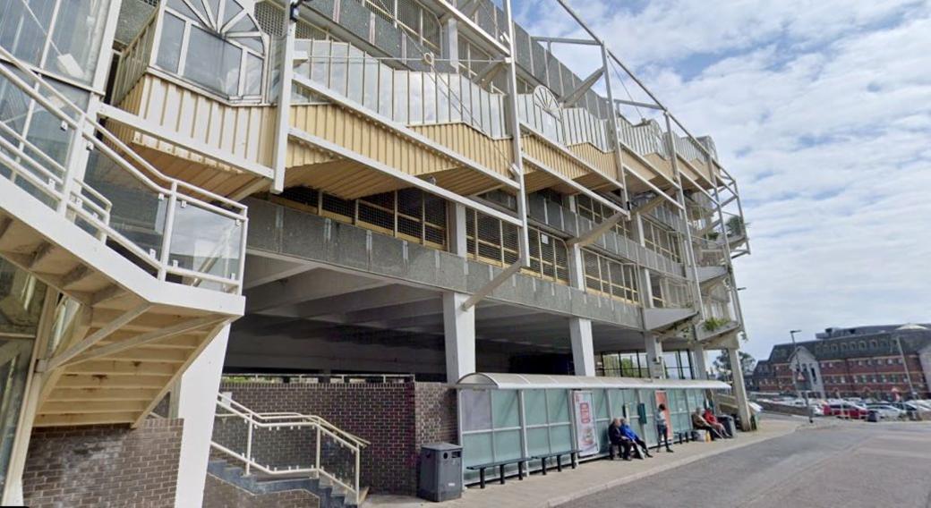 A bus shelter in Sherborne Road in Newton Abbot. It shows a multi-storey car park with a stairway on the side of the building. Below is the us shelter with people waiting for a bus.