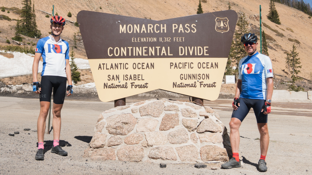 Joshua and George, both in blue and white cycling gear with RBL and a red poppy logo on their tops, stand beside a brown and cream sign which reads: Monarch Pass Elevation 11,312 feet Continental Divide, Atlantic Ocean, San Isabel National Forest, Pacific Ocean Gunnison National Forest 