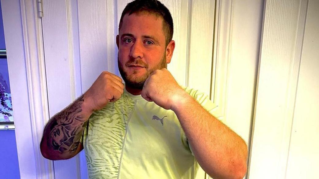 Photo of Dan Mitchell standing in his home wearing a yellow sport T-shirt. He has his fists raised and clenched in a fighting pose.