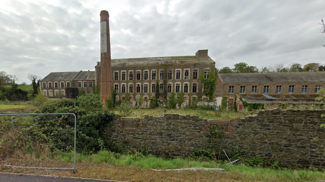 The former Sinton's Mill pictured in April 2022.  The complex is a mixture of brick Victorian buildlings of different heights surrounding a red brick chimney stack.  Many of the windows are broken and there is an uneven wall surrounding the site. 