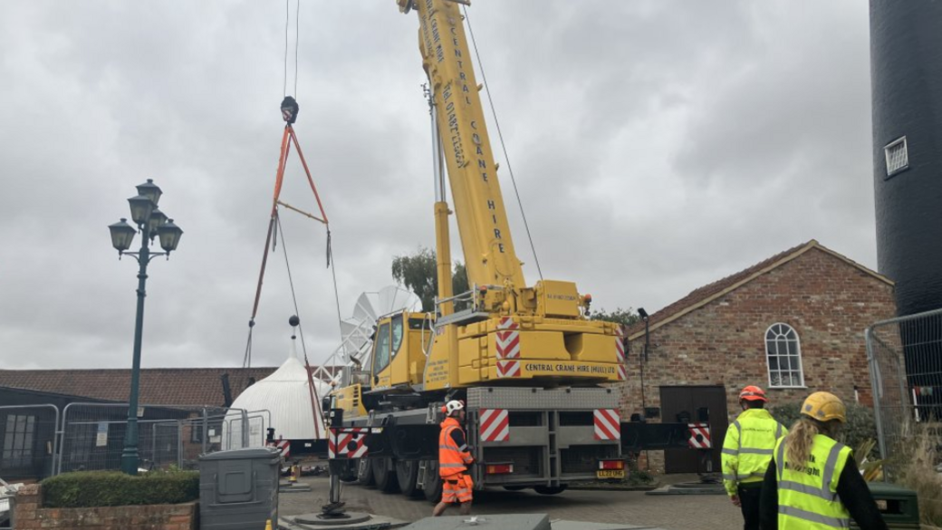 A large yellow crane is starting to lift a white wooden cap onto the top of a windmill. the cap is dome shaped. There are three people wearing high vis vests and hard hats standing near the crane. The sky is grey