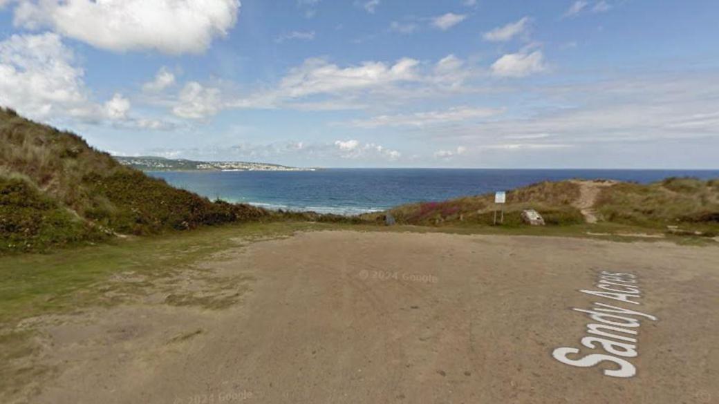 A street view of Sandy Acres coastal car park. The car parking area is brown and is surrounded by green grass and hills. In the distance is blue sea. 