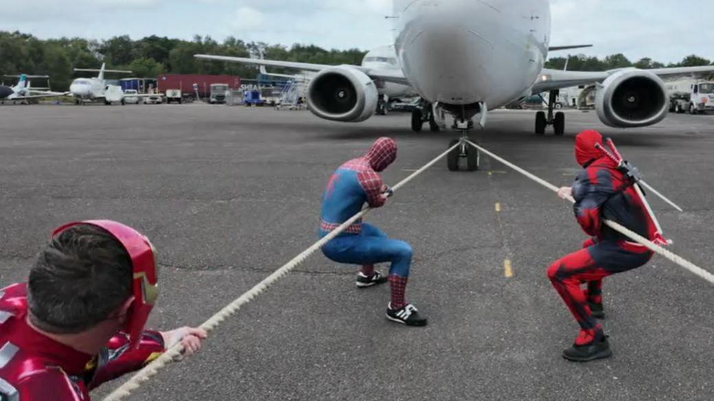 Three men dressed up as superheroes Iron Man, Spiderman and DeadPool are pulling a plane by two ropes