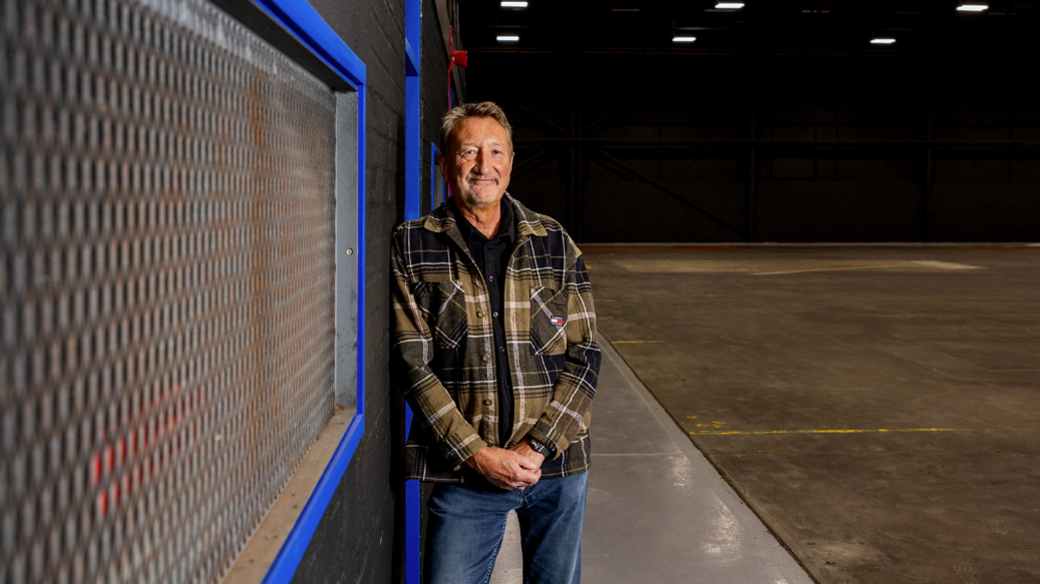 Stephen Knight is wearing blue jeans and a brown checked jacket. He is leaning against a wall