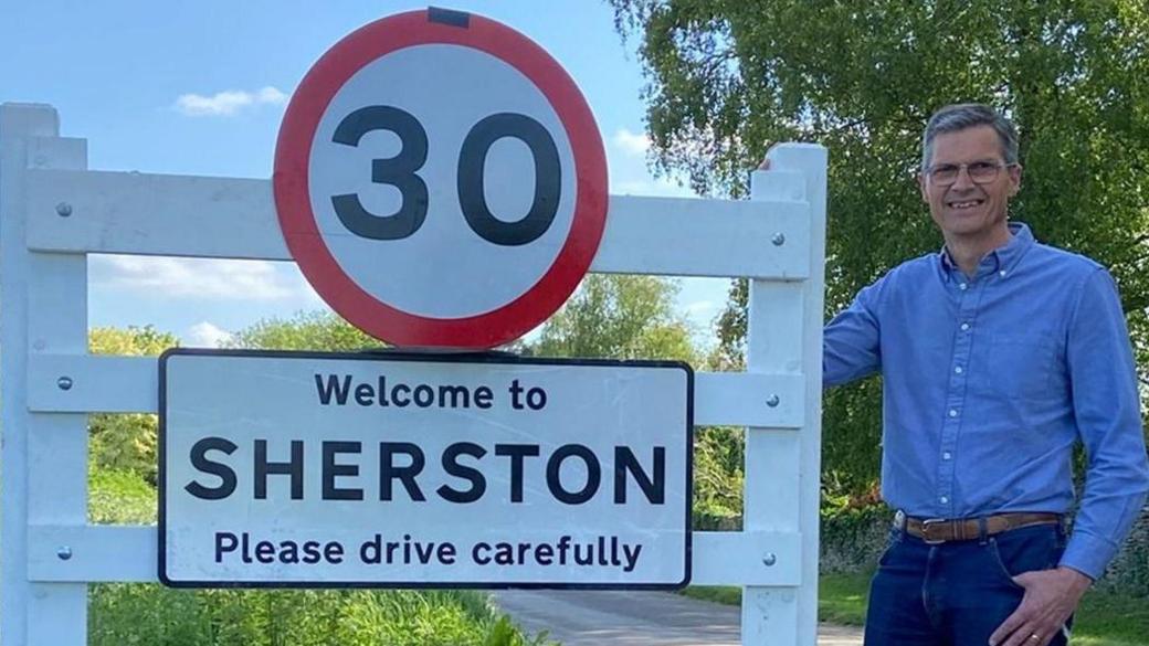 A man in a blue shirt, Wiltshire Cllr Martin Smith, stands beside a 30mph speed sign placed above a 'Welcome to Sherston village' sign. The road is rural with greenery around it.