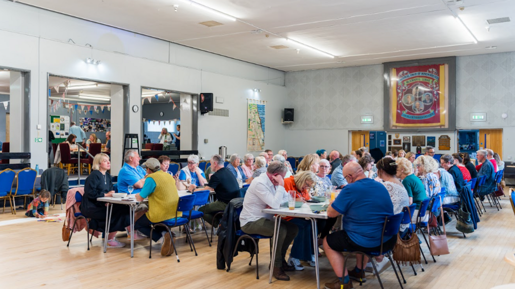 About 50 people are sitting around two rows of tables at Blackhall Community Centre during a listening party for the record. A red banner and a map are hanging on the walls of the brightly-lit room.