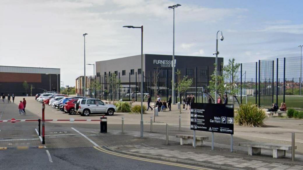 Furness College. A grey cube building stands behind a number of football pitches on the college campus. There are parked cars and people milling about. A dark grey, large rectangular sign in the foreground shows directions to various buildings. There are two thin red and white pole barriers at the entrance to the parking area.