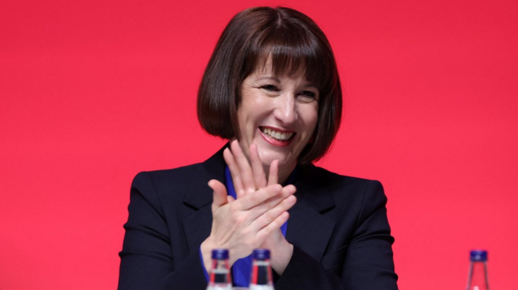 Chancellor Rachel Reeves wears a black suit jacket and a royal blue shirt as she applauds behind a desk at conference