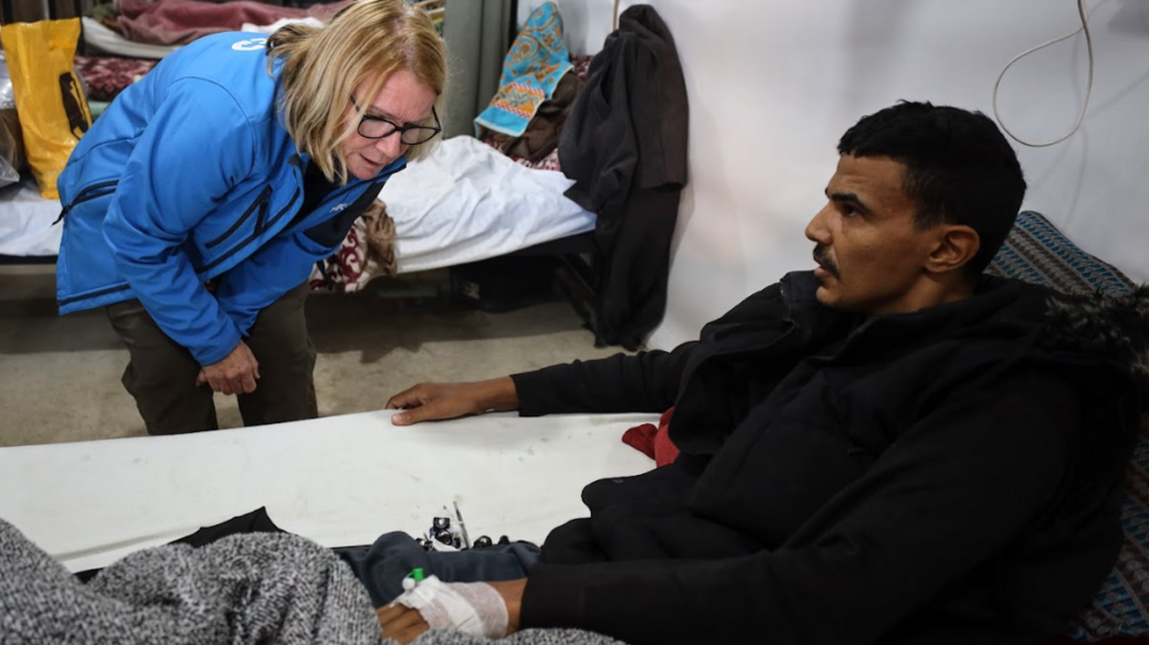 A woman with blonde hair and glasses, wearing a blue jacket, leans in to look at the canular on the back of a man's hand. He is laying on a bed and wearing back clothing