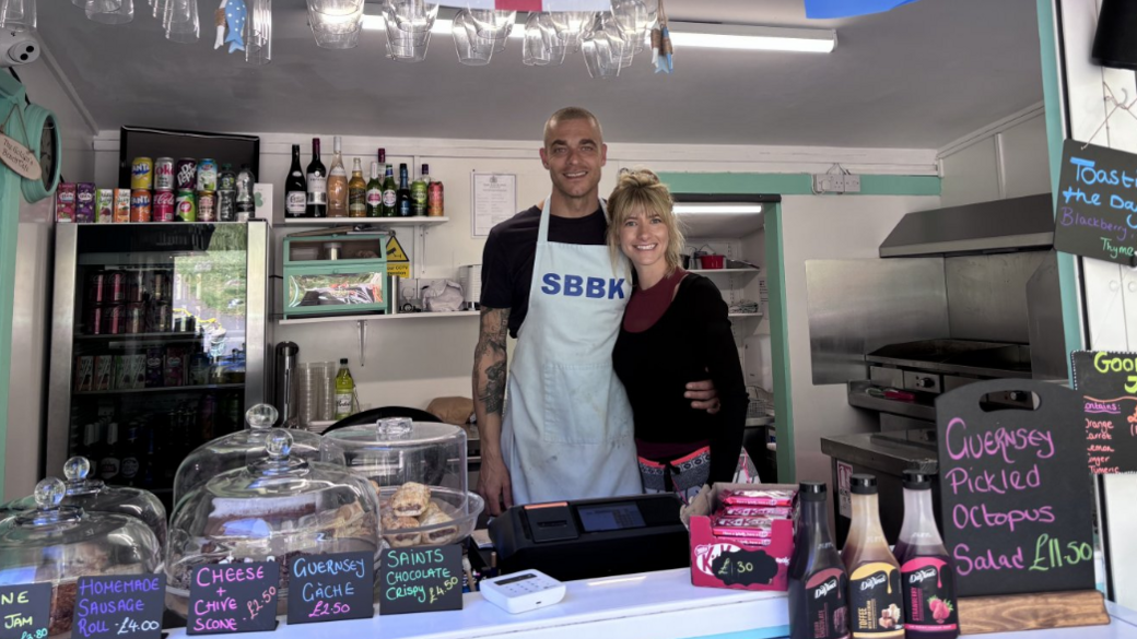 Lucy Jenkins and Antony Safari in the Saints Bay kiosk in Guernsey - they are looking at the camera and smiling. A selection of drinks and food items are on display in the kiosk. Glasses are hanging from a shelf above their heads. 