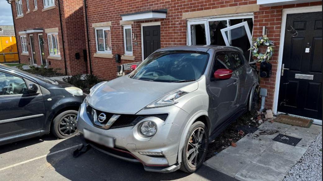 Grey Nissan Juke small hatchback in front of the house, with ground floor front window panels smashed open behind it. Some debris can be seen to the side of the car on the ground, next to the path to the the front door.
