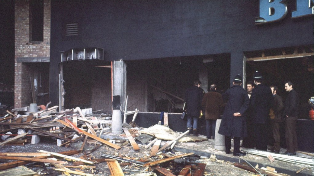 Wreckage including splintered wood and rubble on the pavement outside a building whose front has been blown out. Two policemen in uniform and helmets stand with their backs to camera in a group with three other men in dark clothing. Two men in coats stand looking inside the ruined building with their backs to the camera.