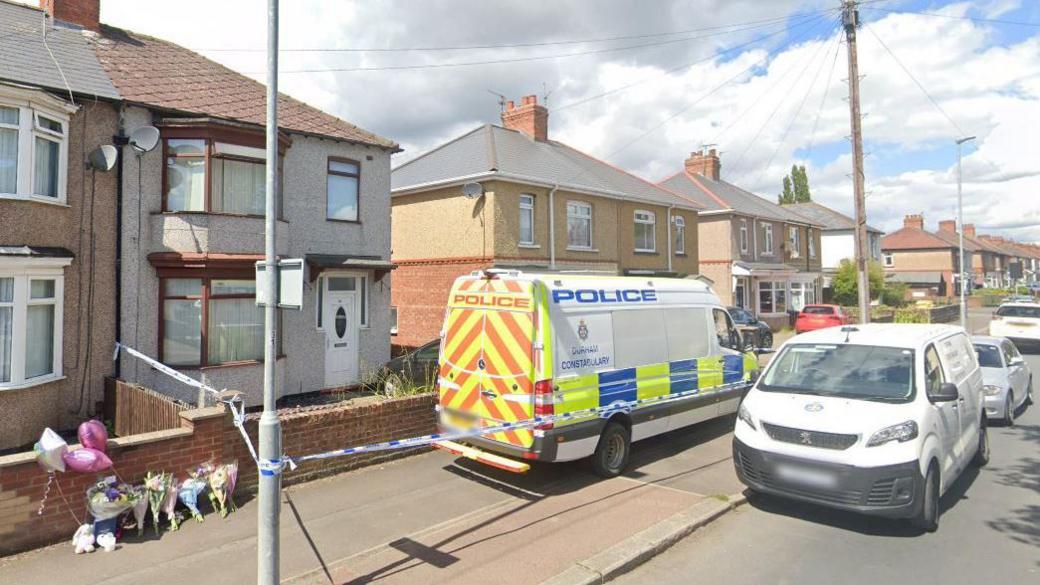 A large police van parked outside a two-storey semi detached house with grey walls, a white door and brown window frames around two large bay windows one above the other. A strip of blue and white police tape runs form the house to a lamppost and above the pavement. Seven bouquets of flowers and several pink balloons are resting against a wall.