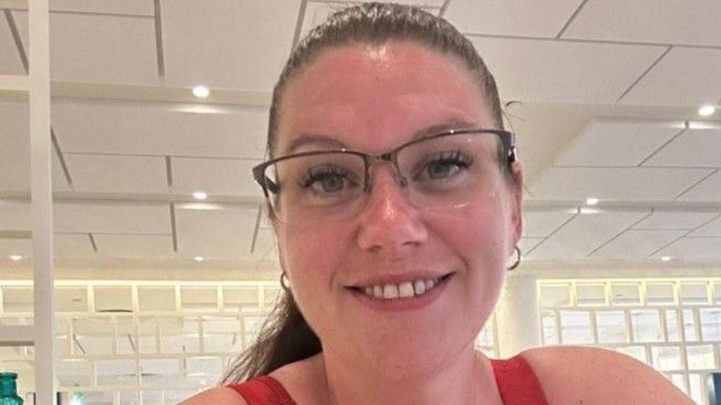 a woman with long brown hair tied back into a ponytail, wearing a red vest top and glasses, smiles at the camera