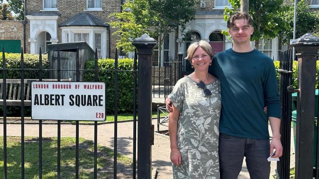Sarah Rozee and her son, who is significantly taller, standing at the fictional Albert Square, with the terraced houses in the background. It is sunny.