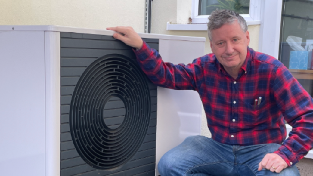Michael bent down next to a piece of equipment he had installed. He is wearing a flannel red and blue shirt and blue jeans.