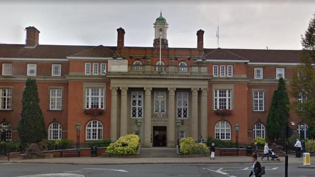 A large brick building with trees and bushes at the front. It lies on the side of a main road. People are walking on the footpath outside.