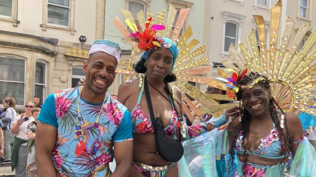 A group shot of three people in bright blue and pink costumes including headdresses. 