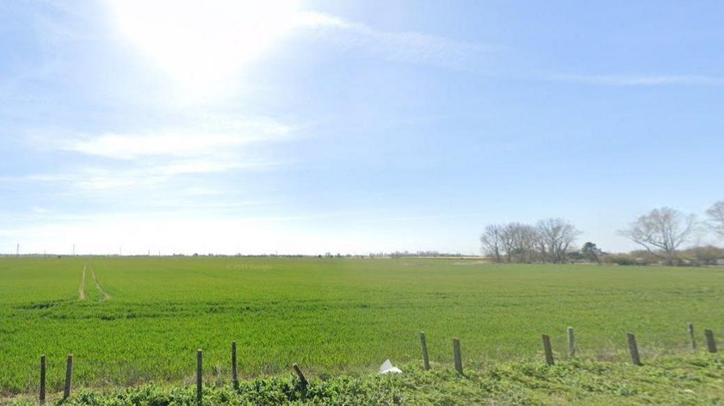 A field stretching uphill is seen from behind a fence at lower ground. The grass is green and the sky is blue with light white clouds and there are bare trees seen at the far edge of the field.