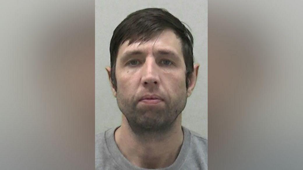 Mugshot of Ormston. He has faint black stubble and thick dark hair swept across his forehead