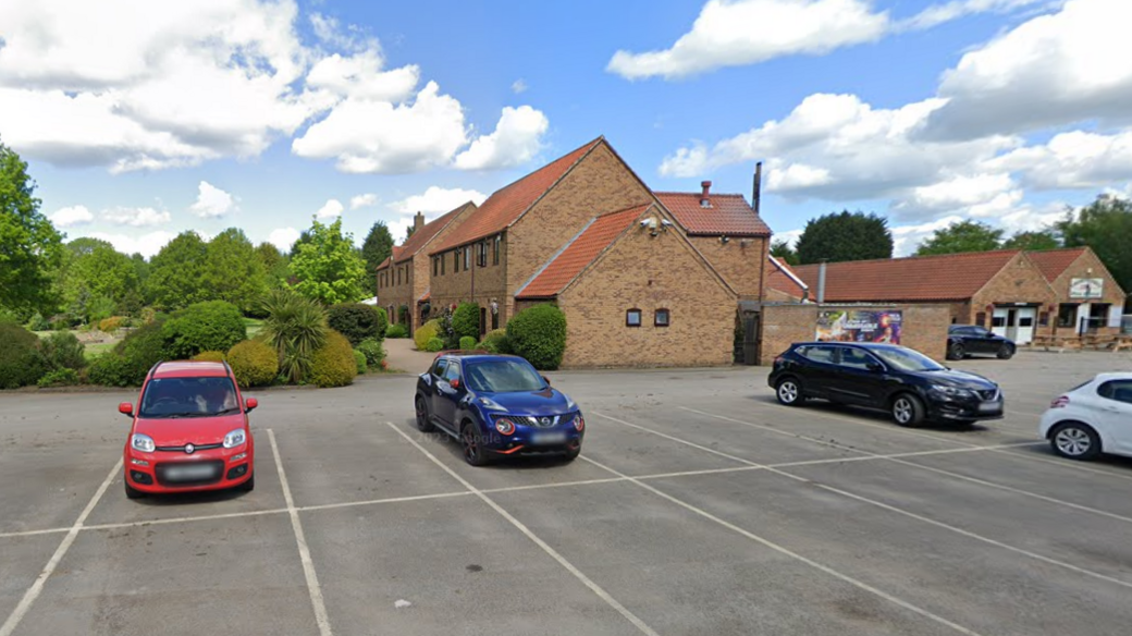 A car park at a country hotel. Pictured are a cluster of parked cars, in amongst empty spaces against the backdrop of some hotel buildings, all of which have sloping rooves and some trees.