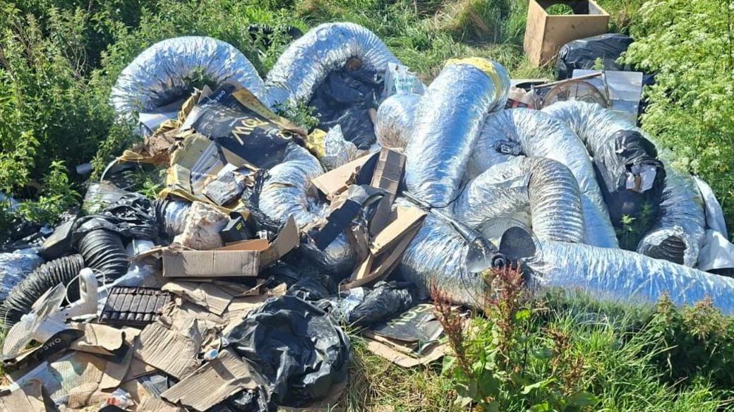 A fly-tip in the countryside with various bits of cardboard and tubes covered in foil used in cannabis factories. Grass and long weeds surround the dumped waste.
