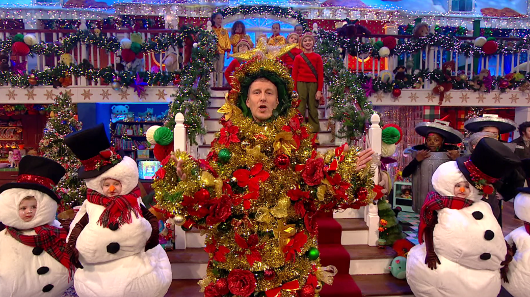 Paddy Kielty covered in Christmas decorations and children dressed as snowmen and others standing on stairs behind him. The stage is also decorated for Christmas