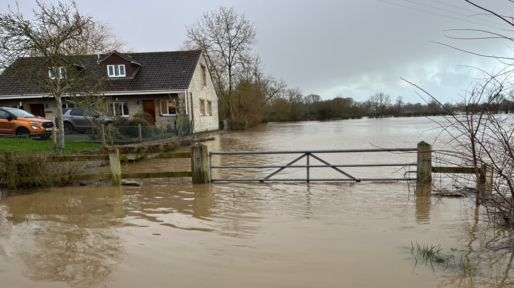 The picture shows a farm gate and land under water. There is a house and two cars to the left of the image.