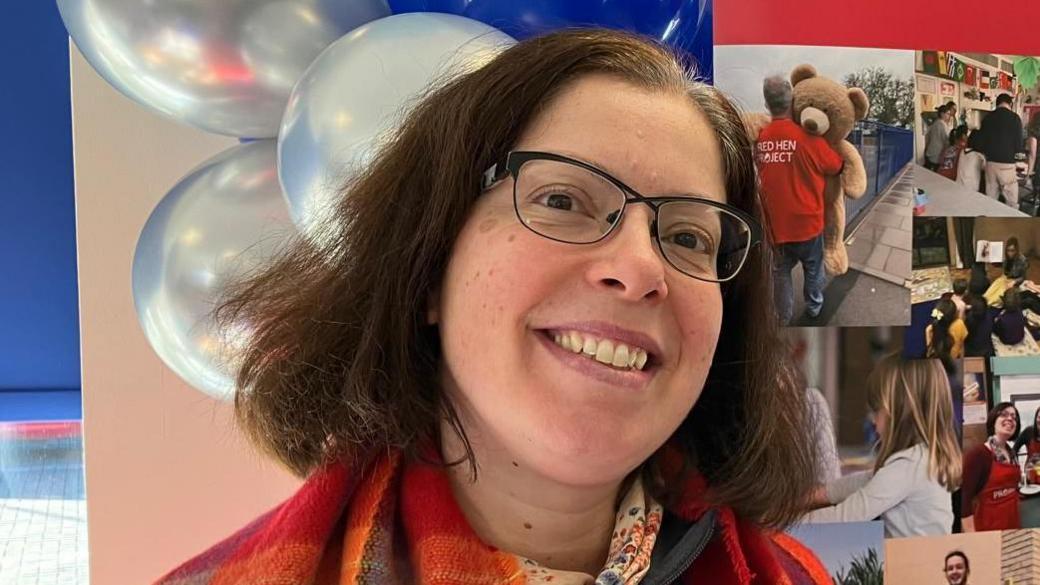 Maria David is looking at the camera, she has a checked red scarf, and is standing in front of the charity's branding, along with photos.