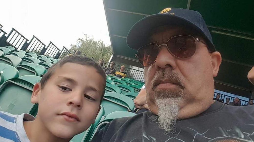 A man and a young boy sit in a sports ground that has rows of green chairs. The man is wearing a baseball cap and sunglasses, and he has a grey goatee beard. The boy has brown hair and he is wearing a blue-and-white striped T-shirt.