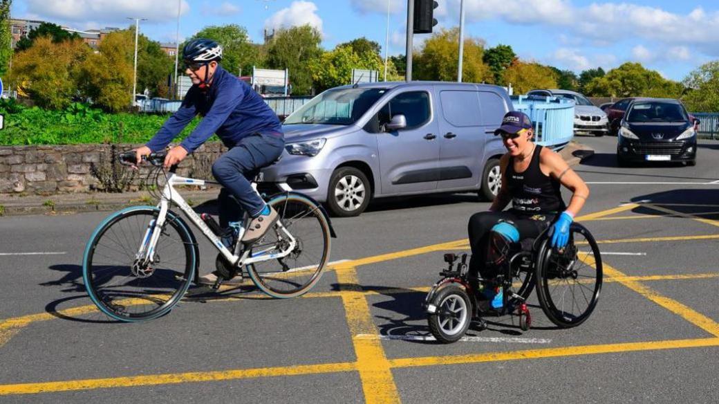 Lexi on her End2End challenge. She's in her wheelchair wearing a hat and blue gloves on roundabout. A bike and cars and a van are around her. 