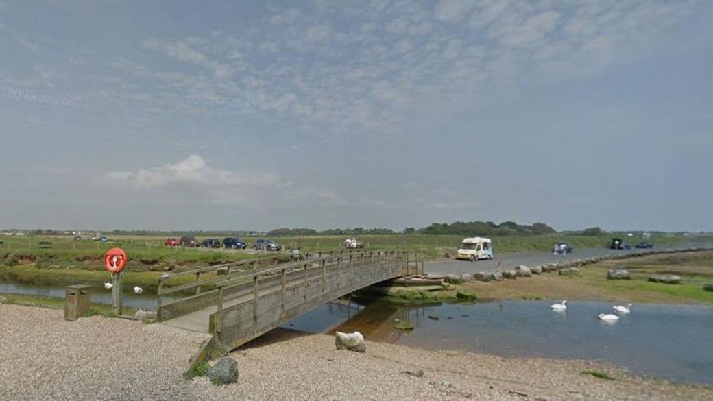 View from the shingle beach towards the road where there are about a dozen vehicles and and an ice-cream van parked. Between the beach and the road is a footbridge and a river where five swans are swimming.
