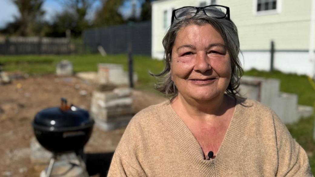 Andrea looks at the camera, she has shoulder length grey hair and her glasses are perched on top of her head. She is wearing a brown jumper. Behind her is an empty caravan pitch, with a barbecue in the background and a static caravan to the right.  
