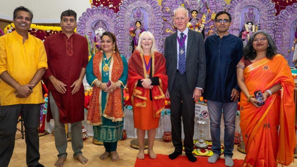 A group of people at an event. Most are wearing saris and Indian clothes but one man is wearing a suit and a chain of office.