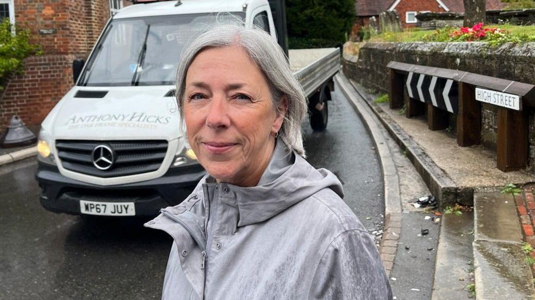 A caucasian woman in her 50's with short white hair smiles at the camera whilst wearing a grey raincoat. A tight bend in the road is visible behind her as is part of a church yard and a white flat bed truck.