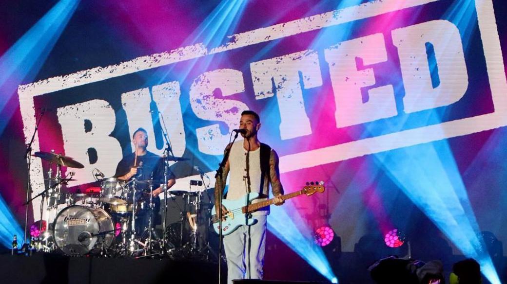 Band members of Busted performing on stage. There is one guitarist and a drummer. Behind them there is a large screen with the band's name.