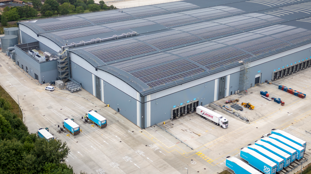A large warehouse with solar panels on its roof surrounded by lorries and trailers.