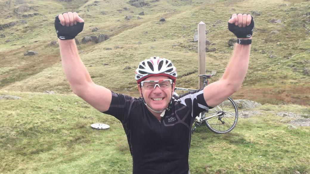 A man in a black cycling top and white cycling helmet with both arms held up in the air and a bicycle behind him on the grass