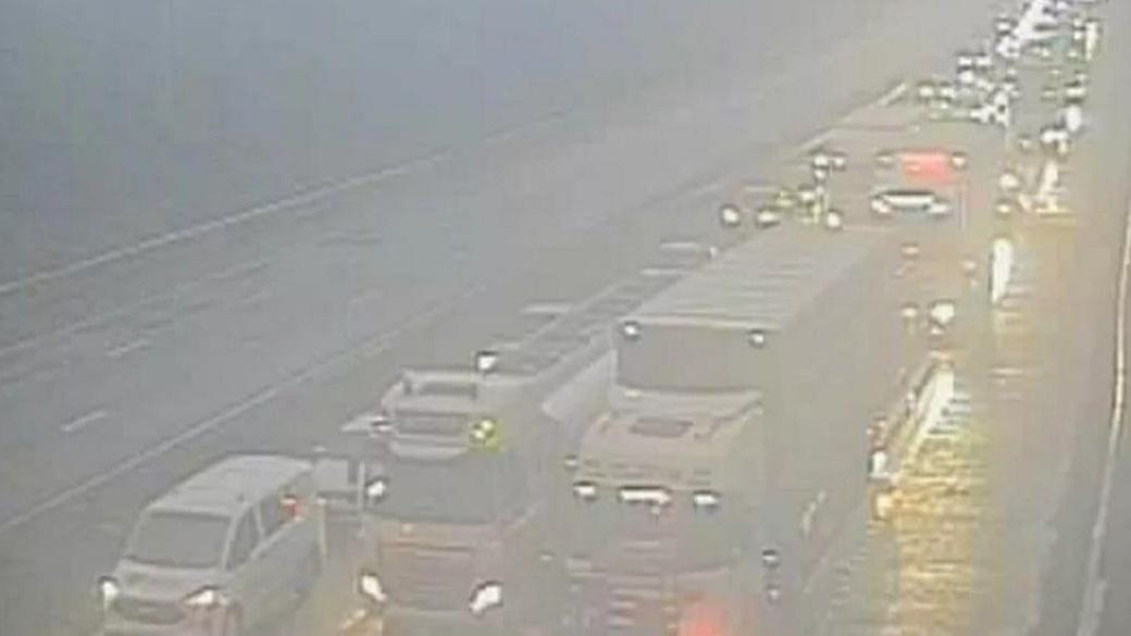 A greyed-out and slightly blurred image from a motorway camera shows a long line of cars, vans and lorries with headlights on queuing into the distance in rainy conditions on one carriageway of the M6, while the other carriageway is free of vehicles