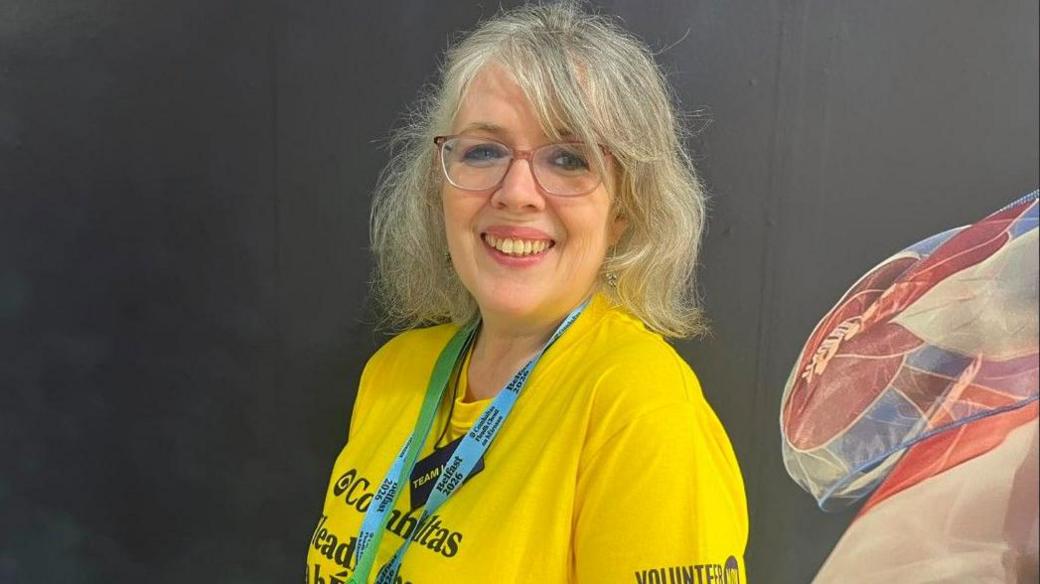 A woman, Camilla Reynolds pictured In front of a black wall with blue, white and red wall art on the right corner.  She has grey hair and is wearing a yellow t-shirt with a blue lanyard and is wearing red glasses. 