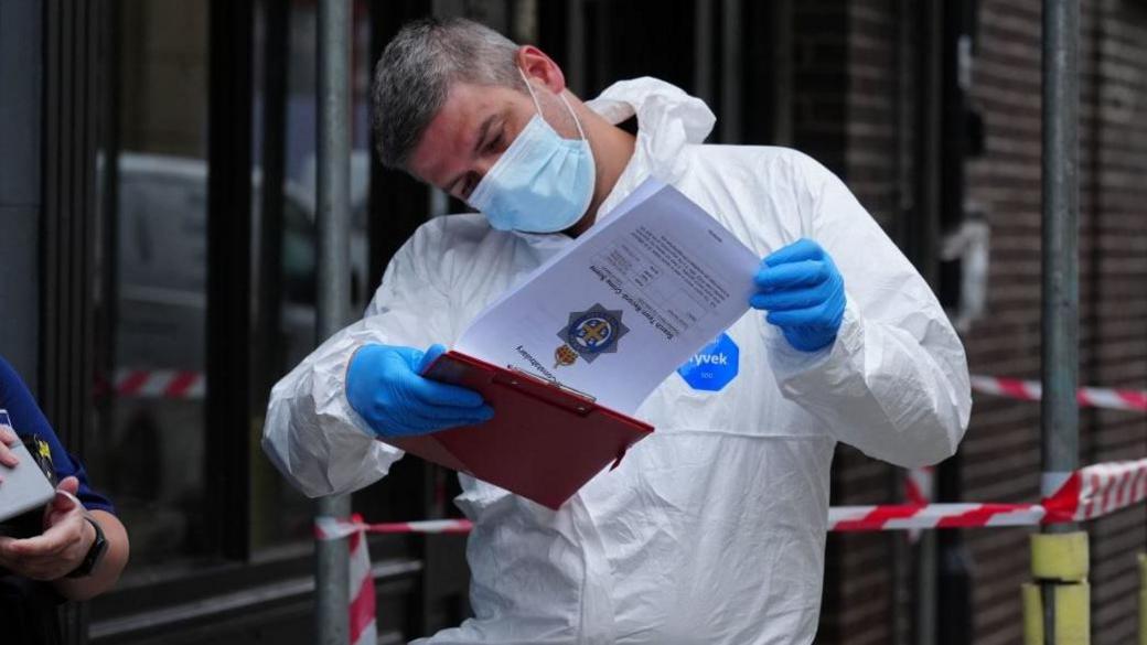 A forensic expert in protective clothes and a mask is outside the cordoned off house looking intently at a clipboard with a paper which has the Durham Police logo