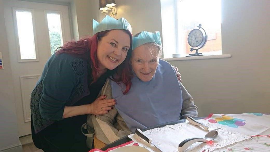 Susie Crozier-Flintham is standing with her hands around her father, Howard Crozier's shoulders. He's an elderly man they are both wearing paper Christmas hats. He's sitting at a table.