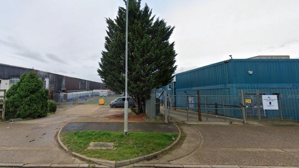 Land on an industrial estate, with a grey building to the left and a blue one to the right. The area between has a large tree, and a car parked on hardstanding. There is another tree in front of the grey building. A sign to the right says "Adrenaline Alley".