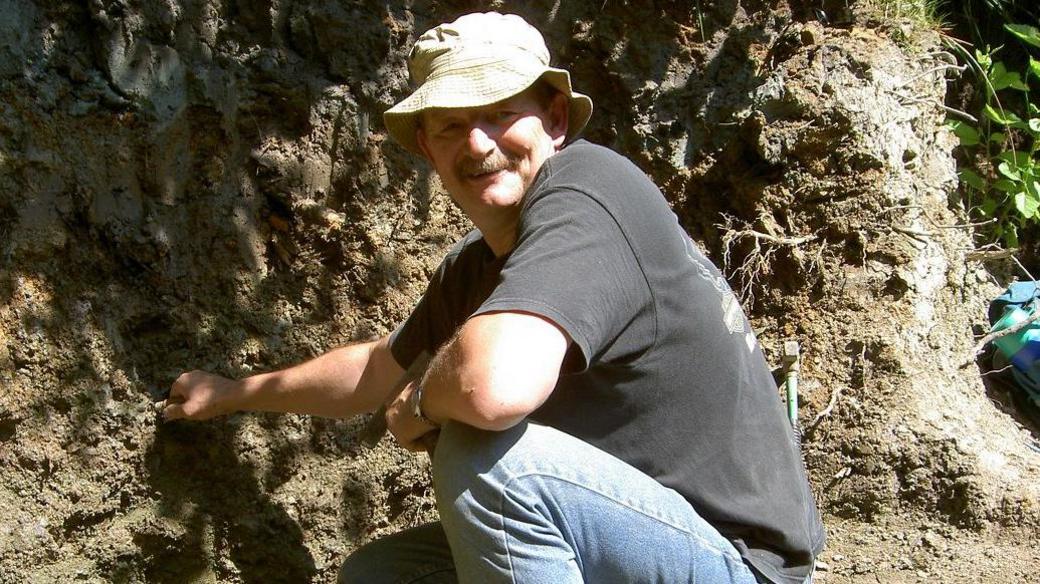 Retired quarryman Dave Brockhurst, wearing a hat, t-shirt and jeans, crouches down to face the camera in the sunshine, at the site where the tyrannosaur and raptor teeth were discovered.