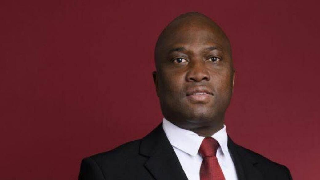 Dominique Désiré Erenon stands in front of a dark red background. He wears a white shirt, red tie, black suit and faces the camera.